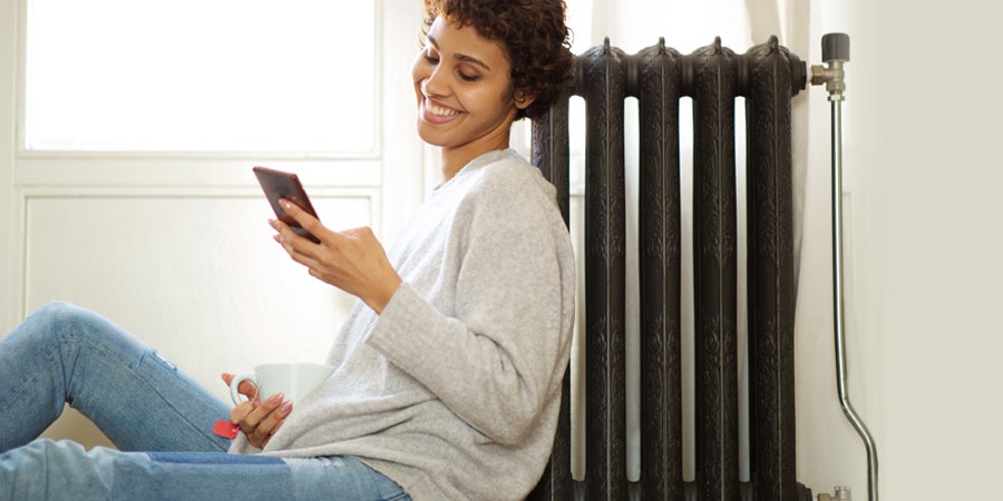 Person sitting on the floor with their back against a black radiator with a big smile across their face. The person has a mug of tea in one hand and their phone in the other hand, searching for an answer to the question 'Do radiator covers block heat?'.