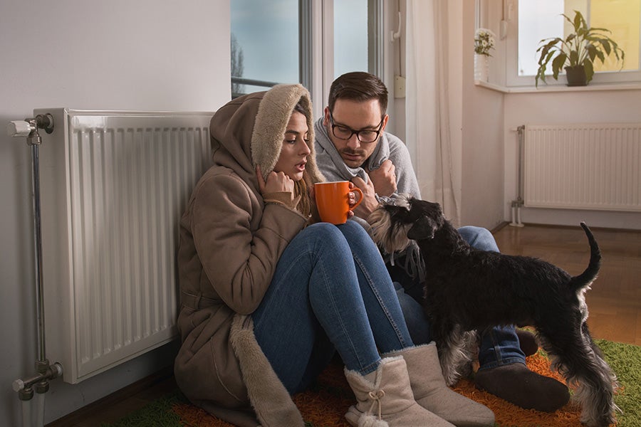 A couple and a dog sitting next to the radiator in their home with no heating or hot water.