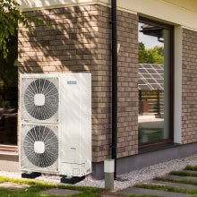 Outdoor heat pump with a sleek, modern design, mounted on a wall. It is set against the backdrop of a contemporary property and a well-maintained garden with greenery.