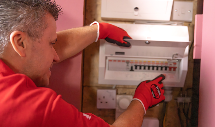Craig Phillips holds open a fuse box, wearing a red t-shirt and gloves.