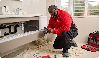 A HomeServe plumber repairing a leak under the bathroom sink for a customer with plumbing and drainage cover.